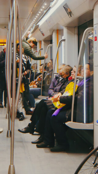 People are seated on a train while a man stands and holds a support bar on public transport.