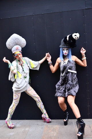 Two women in unique and vibrant fashion outfits pose in front of a dark wall, showcasing creative style.