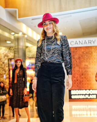 A model showcases a stylish outfit with a pink hat at a fashion show inside a shopping mall.