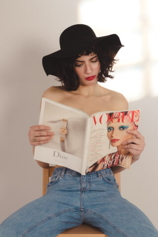 A fashionable woman in a hat and jeans reads Vogue magazine. The image has a soft, editorial style.