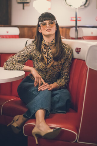 A woman in a leopard print top and heart-shaped sunglasses sits in a retro diner booth.