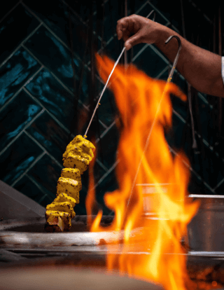 A chef prepares paneer kebab on a grill with fire.