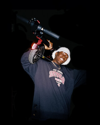 A smiling man in a bucket hat carries an e-scooter at night.