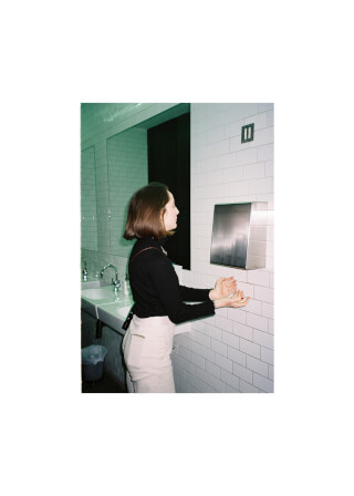 A woman dries her hands in a public restroom with white tile walls and an automatic hand dryer.