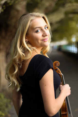 A blonde woman holds her violin, looking over her shoulder in a park.