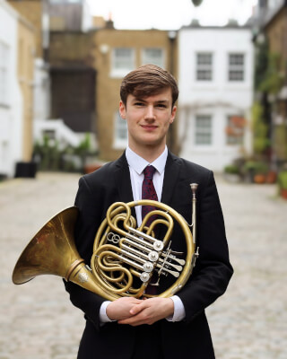 A young man in a suit poses with a French horn in a city courtyard.