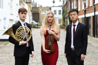 Three young musicians pose outdoors in formal wear with a French horn and violin.