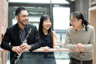 Three musicians are smiling and leaning on a railing inside a building, with a clarinet and cello. Happy and cheerful atmosphere.