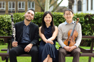 Three smiling musicians sit on a bench outdoors, holding a clarinet and violin.