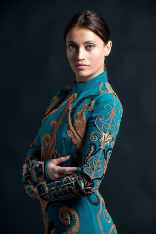 A stylish woman in an embroidered teal dress poses in a studio portrait.