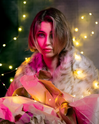 A woman in a fur coat sits amidst wrapping paper and fairy lights in a moody editorial style.