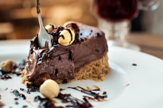 Close-up of a decadent chocolate cake slice with hazelnuts and sauce, partially eaten with a fork on a white plate.