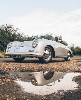A classic silver Porsche Speedster reflects in a puddle on a country road, evoking vintage charm.
