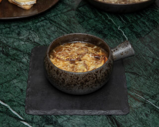 A close-up of french onion soup with melted cheese, presented in a rustic bowl on a black slate.