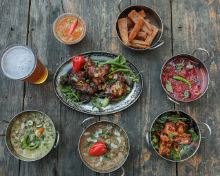 Overhead shot of Indian food including curry, chicken wings, and beer on a rustic wooden table.