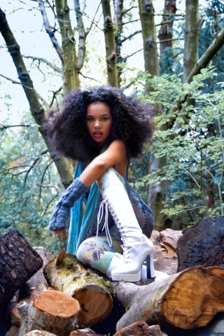 A woman with big curly hair sits on a pile of logs in the woods wearing white boots.
