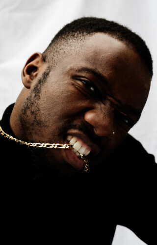 A confident man bites down on a gold chain in a close-up studio shot with intense expression.