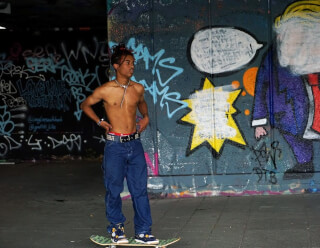 A young shirtless man stands confidently on a skateboard in an urban setting with graffiti art behind him.