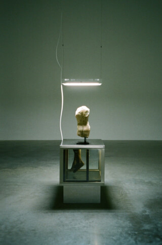 A minimalist art exhibit features a fragmented statue illuminated by a hanging light fixture in a gallery space.