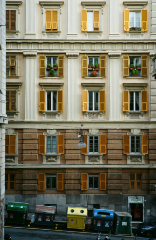 Building facade with yellow shutters on windows, plus trash cans on street level.