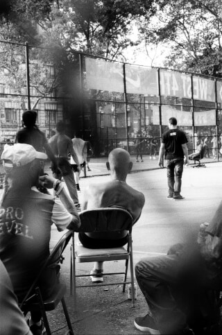 A black and white image captures an urban basketball game with spectators and players in a public park setting.