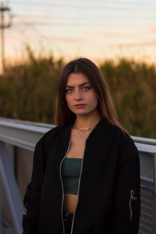 A young woman in a bomber jacket gazes intently at the camera in a natural outdoor setting.