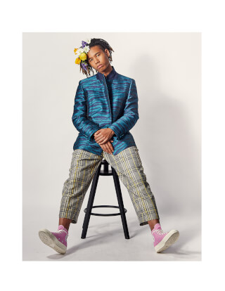 A stylish man with flowers in his hair sits on a stool, showcasing a unique fashion ensemble for editorial or advertising use.