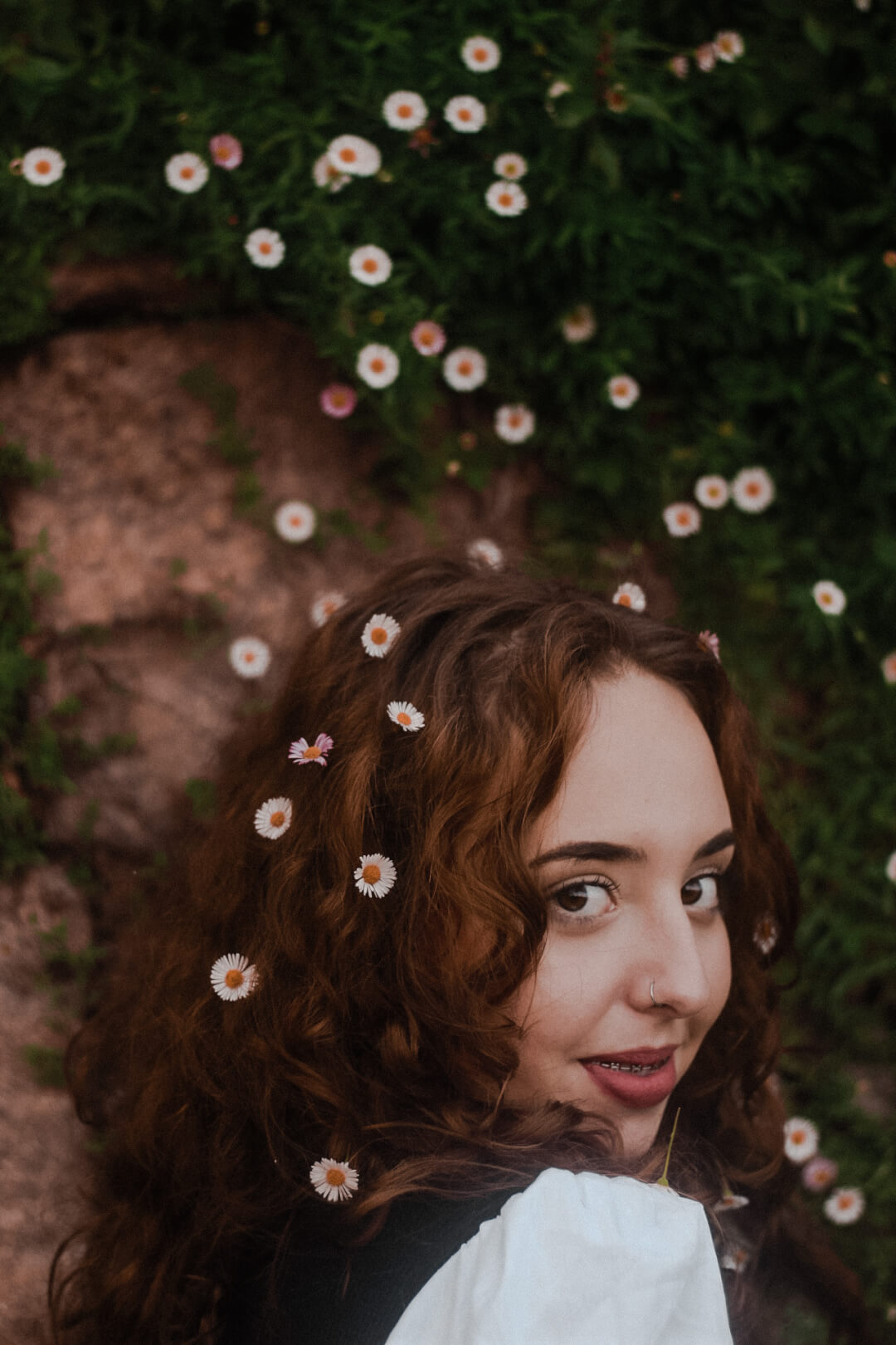 A beautiful girl with daisy flowers in her curly brown hair smiles dreamily in a lush garden.