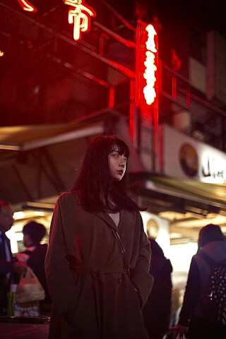A woman stands on a city street at night under the glow of red neon lights. 
