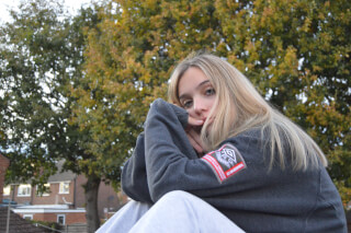 A young woman poses outdoors in a gray sweatshirt with fall foliage behind her.