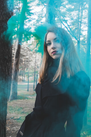A woman in a black coat stands in a forest surrounded by blue smoke.