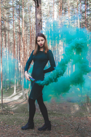 A woman in a dress stands in a forest holding a smoke grenade with vibrant teal smoke swirling around her.