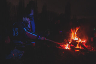 A man smokes by a blazing campfire at night, surrounded by friends at an outdoor gathering.