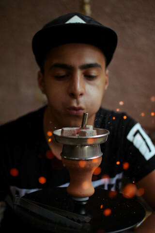A young man blows on hookah coals, captured with shallow depth of field in an intimate and relaxing indoor setting.
