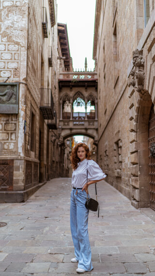 A redhead woman in jeans stands in a historic alleyway, blending fashion with urban exploration.