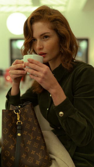 A red-haired woman in a green shirt sips from a coffee cup with a Louis Vuitton handbag nearby.