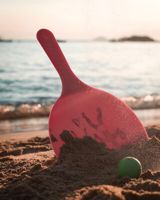 A red beach paddle and green ball sit in the sand on a sunny beach.