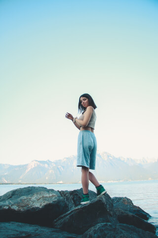 A young woman poses on rocks by a lake with mountains in the background on a sunny day.