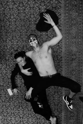 A man in a skull mask and a woman are lying on a carpet.