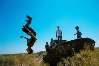 A boy flips in front of a rusty boat where other children are playing against a clear blue sky in a desert landscape.