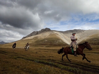Three men ride horses across a grassy field, a mountain rising behind them on an overcast day.