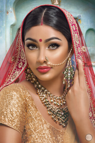 A glamorous Indian bride in traditional attire, adorned with gold jewelry and a red veil, poses elegantly for a close-up portrait.