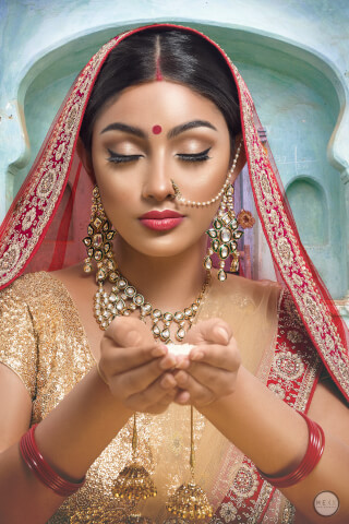 An elegant Indian bride with a closed-eye makeup look, adorned in traditional jewelry and clothing, holding rice grains in her hands.