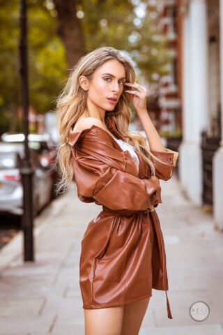 A woman in a brown leather dress poses confidently in an urban setting for a fashion editorial.