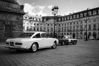 Two classic Lancia Fulvia cars are parked on a cobblestone square in a European city.