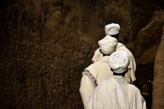 Three religious figures in Lalibela, Ethiopia, wearing turbans and robes stand near a rock formation.