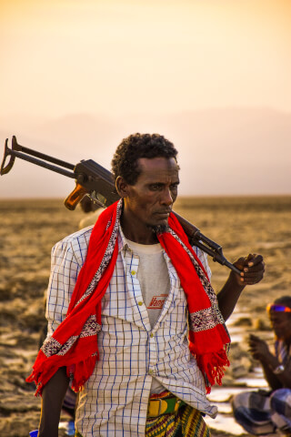 An Ethiopian man in an arid landscape carries a rifle while wearing a red scarf.