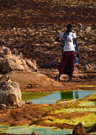 A man stands near a small, green lake in the Danakil Depression, surrounded by colorful mineral formations.