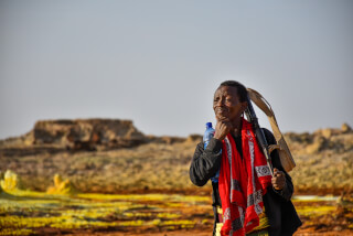 A man with a rifle and water bottle stands thoughtfully in a desert landscape.