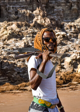 A man with a rifle and sunglasses stands outdoors, exuding confidence.